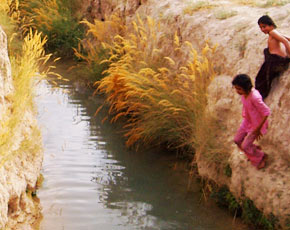 Two young children playing on the earthen bank of a flowing traditional Karez water channel in rural Balochistan