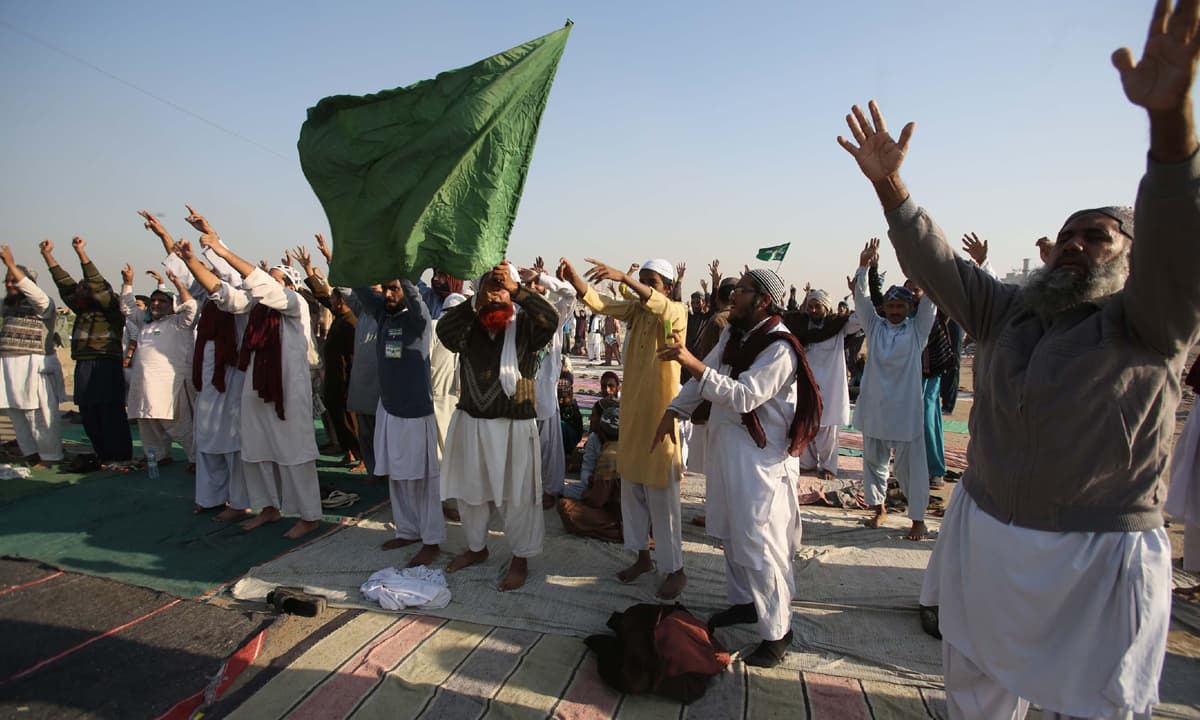 A group of men raising their hands and waving a green flag during an outdoor religious political protest in Pakistan.
