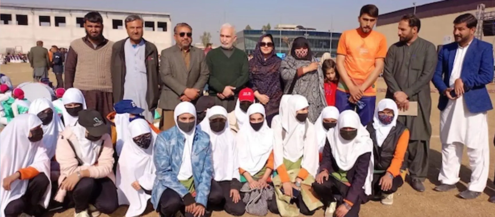 Young female sports players wearing white headscarves and face masks sitting on grass alongside male and female coaches in Khyber Pakhtunkhwa.