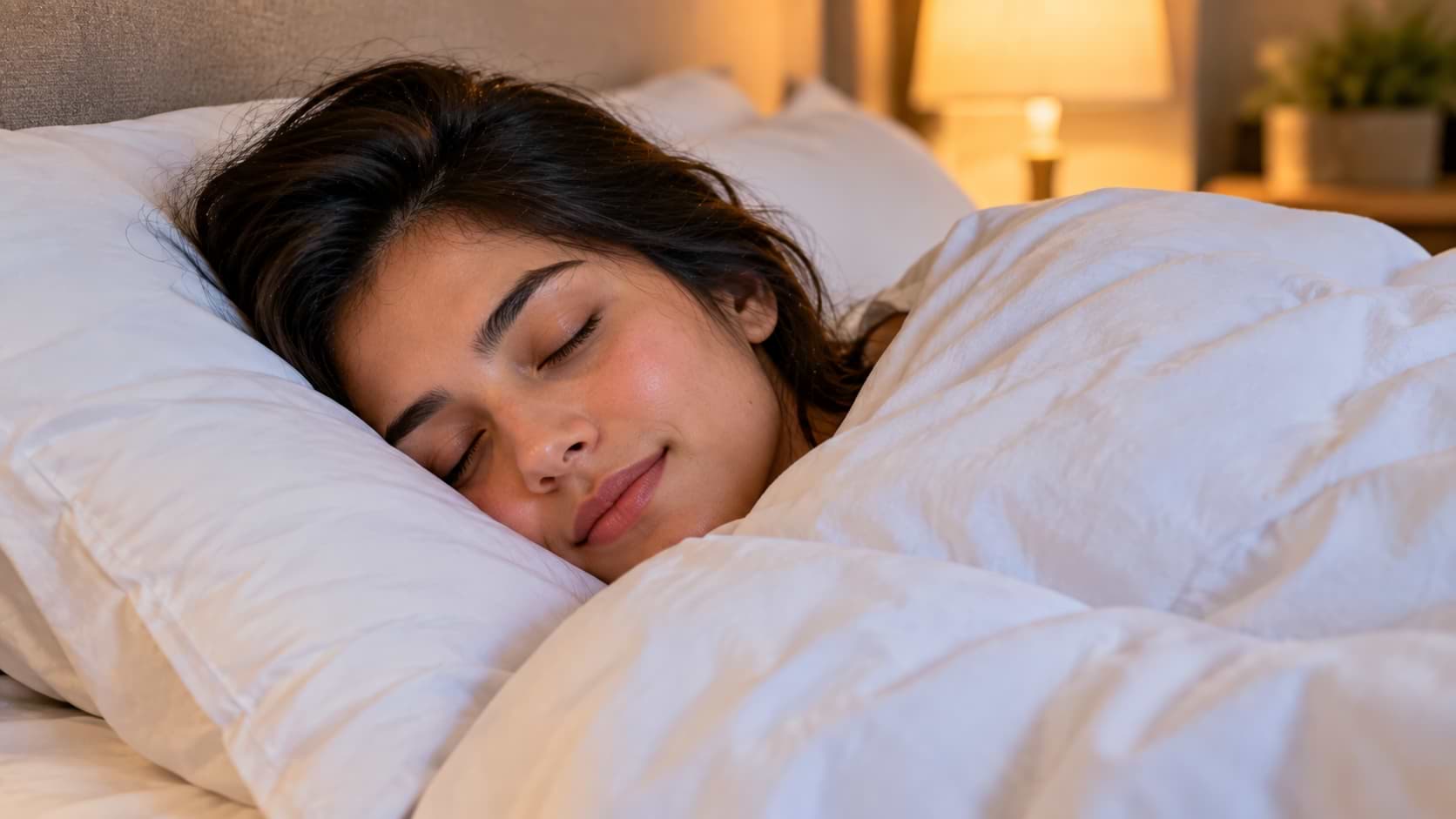 A young woman sleeping peacefully wrapped in a soft white blanket with her head resting on plush white pillows in a cozy bedroom setting.