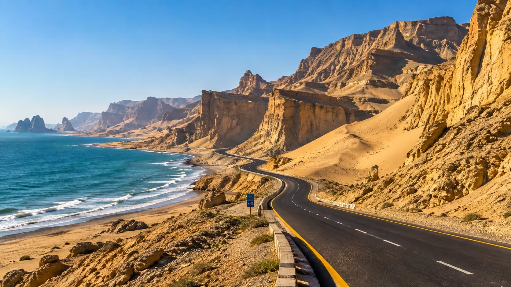 A paved two-lane highway with an N-10 road marker winding between golden desert rock cliffs and the turquoise ocean waves of the Arabian Sea in Balochistan, Pakistan.