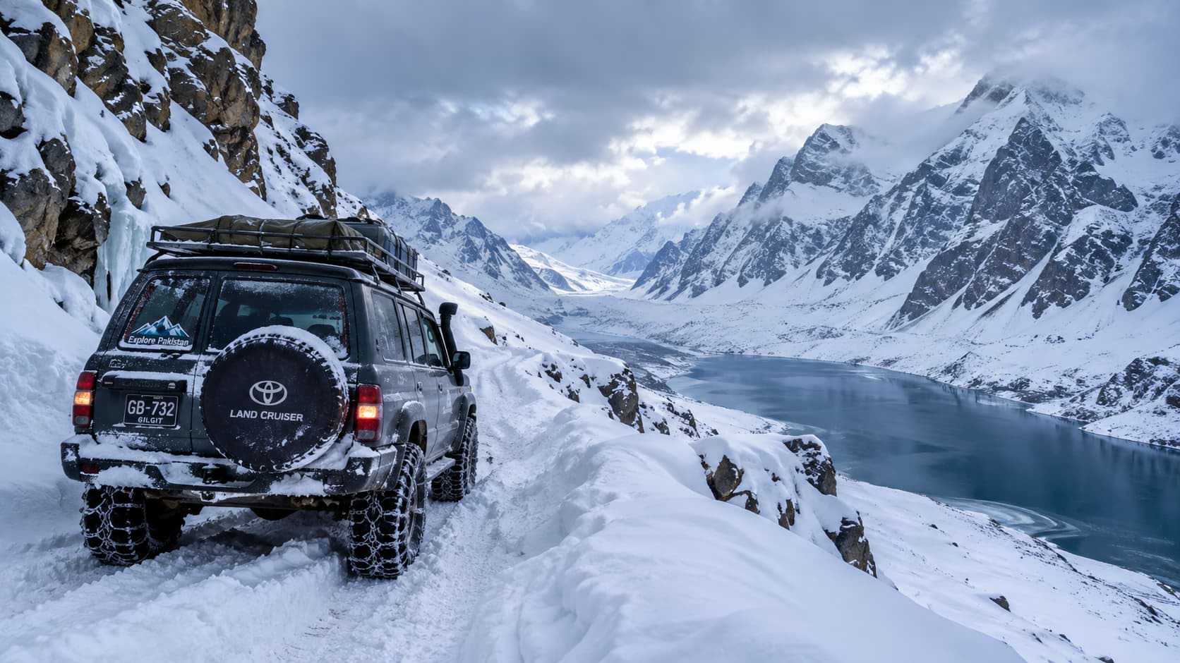 A dark gray Toyota Land Cruiser with tire chains driving on a narrow snow-covered cliffside track overlooking a deep blue mountain lake surrounded by snow-capped peaks in Gilgit-Baltistan, Pakistan.