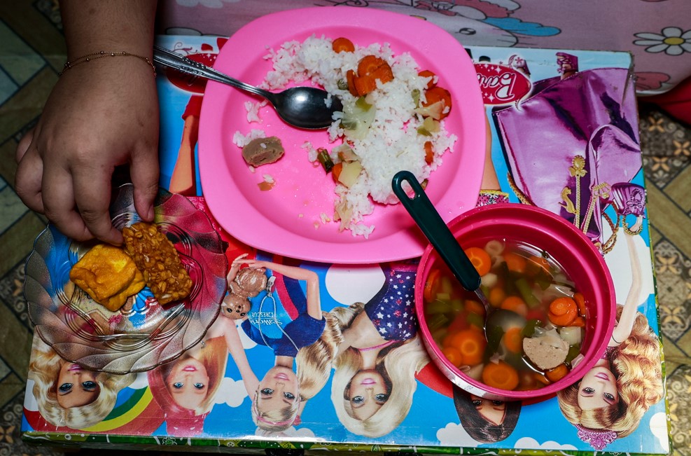 A top-down view of a hand reaching for fried tempeh next to a pink plate of white rice with vegetables and a pink bowl of carrot soup on a colorful table.